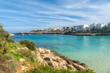 Turistler, güneşli bir günde arka planda beyaz evler ve teknelerle Mallorca, İspanya 'da kristal berrak turkuaz sularının keyfini çıkarıyorlar.