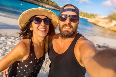 Bir adam ve bir kadın kumsalda kameraya gülümsüyor. Kadın hasır şapka giyerken adam siyah bir atlet ve güneş gözlüğü takıyor.