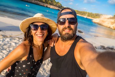 Bir adam ve bir kadın sahilde fotoğraf için poz veriyorlar. Kadın hasır şapka ve güneş gözlüğü takarken adam siyah bir atlet ve güneş gözlüğü takıyor. İkisinin de yüzünde gülümseme var.