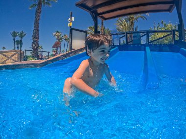 Mutlu çocuk canlı bir su parkındaki parlak mavi bir havuza kayıyor, yaz tatilinin heyecanıyla eğleniyor.