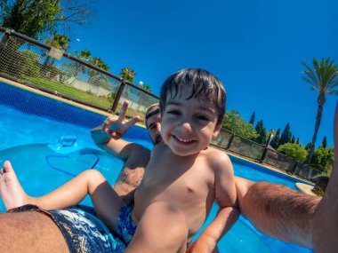 Güneşli bir günde, bir su parkında, canlı yeşillikler ve açık mavi gökyüzüyle çevrili bir havuzda yüzerken gülümseyen genç çocuk.