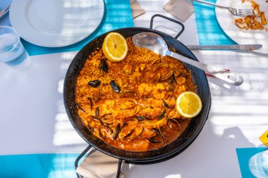 İçinde midyeler, karidesler, istiridyeler ve limon dilimleri bulunan buharlı deniz ürünleri paellası canlı mavi bir masada geleneksel bir tavada servis edilir.