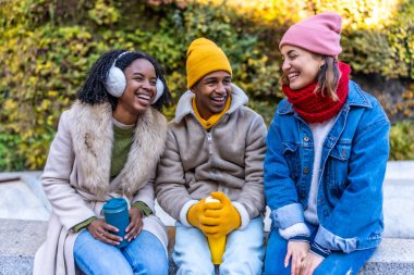 Üç neşeli çoklu etnik arkadaş soğuk bir kış gününde şehirde oturmuş yeniden kullanılabilir kahve fincanlarının keyfini çıkarıyorlar.