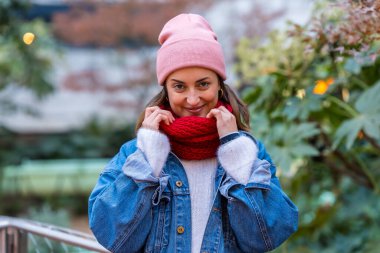 Genç bir kadın şehirde kış gününün tadını çıkarıyor, rahat pembe bir bere takıyor ve canlı kırmızı bir eşarp takıyor, mutluluk ve tarz saçıyor.