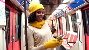 Akıllı telefon ve tırabzan kullanırken gülümseyen Afro-Amerikalı genç bir kadın. Şehir içi iş çıkışı metro istasyonunda duruyor.