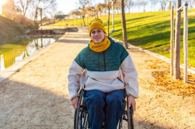 Parkta güneşli bir günün tadını çıkaran tekerlekli sandalyede gülümseyen ve kameraya bakan genç bir adamın portresi.