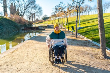 Happy young woman using wheelchair enjoying sunny day in park near a small lake, promoting accessible recreation and inclusivity clipart