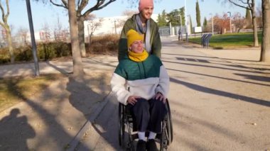 Sevgili çift açık havada eğleniyor. Adam tekerlekli sandalyeyi dikkatlice güneşli parkta itiyor. Bakım, destek ve mutluluğu paylaşıyor.