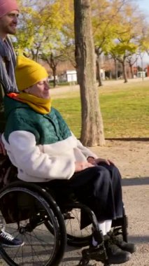 Dışarıda gelişen destekleyici dostluk, tekerlekli sandalyeyi iten genç adam arkadaşını güneş ışığı parkında kullanıyor, neşe ve bağlantı anlarını paylaşıyor.