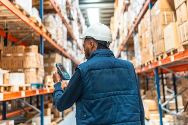 Büyük bir dağıtım deposunda envanter çıkarmak için tarayıcı kullanan Afrikalı genç bir işçinin arka görüntüsü.
