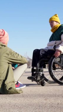 Tekerlekli sandalye kullanıcısı ve arkadaşı kentsel ortamdaki güneşli açık hava anlarında anlamlı bir sohbete girişiyor, iletişimi ve anlayışı paylaşıyor