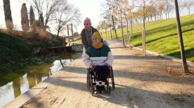Destekleyici arkadaş tekerlekli sandalyeyi manzaralı kanal boyunca iterek güneşli park ortamında eğlenceli anları paylaşıyor.