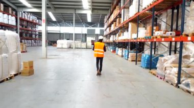 Turuncu yelek ve beyaz kask giyen bir lojistik operatörü depoya giriyor ve malları kontrol ediyor.