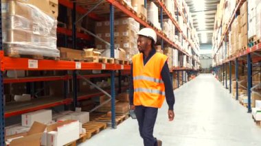 Güvenlik yeleği ve kask giyen bir depo işçisi büyük dağıtım merkezinin koridorlarında yürüyor, mal ve malzemeleri kontrol ediyor.