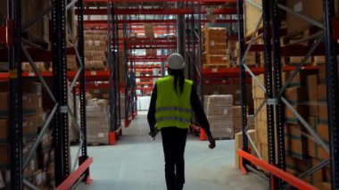 Güvenlik yeleği ve kask giyen bir lojistik operatörü raflarla dolu geniş bir depoda paketleri tarıyor.