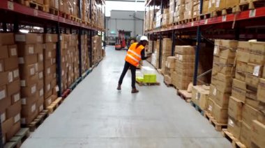 Lojistik işçisi, büyük bir depoda karton kutularla dolu rafların arasından kriko çekiyor.