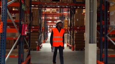 Güvenlik yeleği ve kask giyen bir depo işçisi koridorlarda yürüyor, mal ve envanteri kontrol ediyor.
