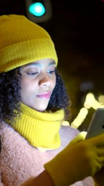 Gece şehir caddesinde parlak sarı kış kıyafeti giyen genç bir kadın bulanık ışıklarla aydınlatılmış, odaklanmış bir ifadeyle akıllı telefona göz atıyor.