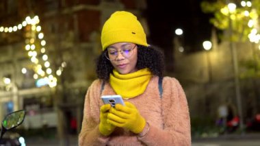 Neşeli, Afro-Amerikalı kadın canlı, sarı kışlık giysiler giyiyordu. Gece vakti şehrin aydınlık sokaklarında dikilirken akıllı telefondan mesajlaşıyordu.