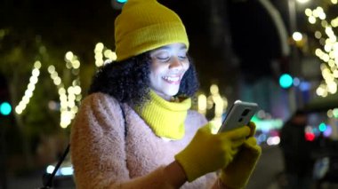 Gece şehir caddesinde akıllı telefon kullanan, yılbaşı ışıklarıyla aydınlatılmış, kışlık giysiler giyen genç bir kadın.