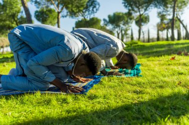 Two senegalese men are kneeling on prayer rugs in a park, performing salat during the holy month of ramadan, dressed in traditional clothing clipart