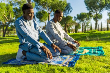 Two young senegalese men wearing traditional clothing are kneeling on prayer rugs in a park, observing ramadan clipart