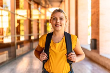 Kampüste kameraya gülümseyen sarışın üniversite öğrencisinin portresi.