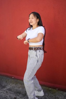 Kentte kırmızı bir duvarın yanında dans eden ve kollarını daire şeklinde hareket ettiren Asyalı genç bir kadının dikey yan görüntüsü.