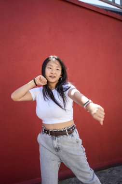 Kırmızı şehir duvarının yanında dans eden Asyalı genç bir kadının dikey fotoğrafı.