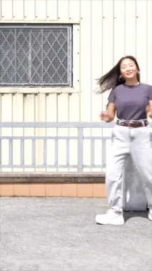 Endüstriyel bir alanda enerjik bir şekilde zıplayan genç ve mutlu bir Koreli kadının dikey videosu.