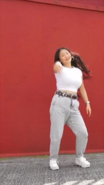 Kırmızı şehir duvarının yanında dans eden Asyalı genç bir kadının dikey videosu.