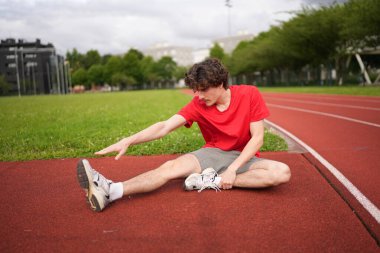 Açık havada koşu pistinde ısınan bacaklarını ısıtan beyaz, genç bir adamın tam boy fotoğrafı.
