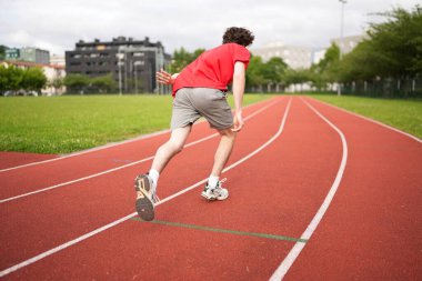 Beyaz tenli genç bir sporcunun koşu pistinde başlangıç çizgisini terk edişinin tam boy dikiz görüntüsü