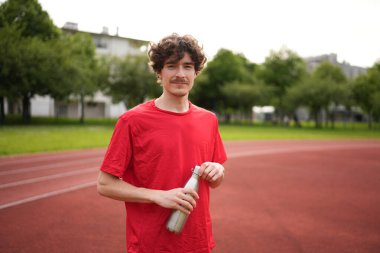 Portrait of a male caucasian young runner with reusable bottle of water after training in an outdoor running track clipart