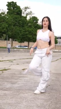 Beyaz tenli genç bir kadının şehir parkında serbest dans edişinin dikey videosu.
