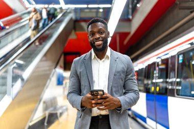 Genç siyahi işadamı istasyonda tren beklerken akıllı telefon kullanıyor. Yürüyen merdiven ve arka planda tren var.