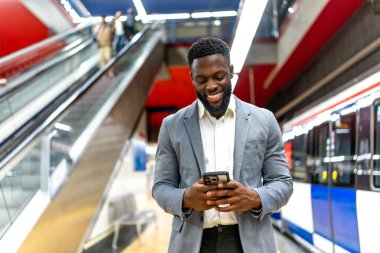 Bir metro istasyonunda tren beklerken akıllı telefon kullanan gülümseyen bir işadamı, işe gidiş gelişi sırasında bağlantı ve rahatlığın tadını çıkarıyor.