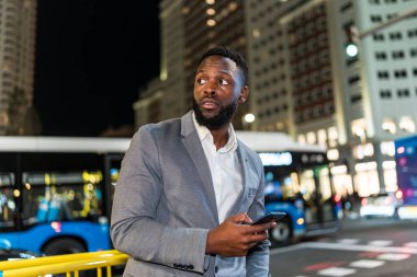 Akıllı bir telefon tutan genç bir işadamı, parlak ışıklar ve modern gökdelenlerle çevrili, hareketli bir şehre gece bakıyor.