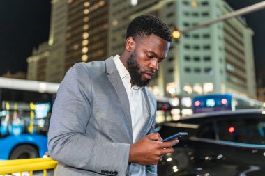 Gece şehir caddesinde akıllı telefon kullanan genç işadamı. Arka planda bulanık binalar ve trafik var.