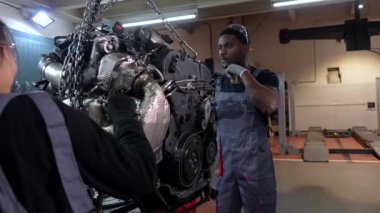 Yoğun bir garaj ortamında iki tamirci büyük bir araba motoru üzerinde çalışıyor.