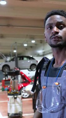 Koruyucu gözlük takan ve gülümseyen bir tamirci bir araba atölyesinde duruyor, uzmanlık ve profesyonellik sergiliyor.