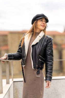 Şehir ortamında poz veren şık bir kadın. Koyun derisi yakalı, siyah deri ceketli ve gazeteci şapkalı.