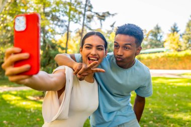 Çok ırklı çift selfie çekiyor zafer işareti yapıyorlar ve parkta öpüşüyorlar.