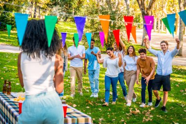 Çok ırklı arkadaşlar parktaki sürpriz bir doğum günü partisine geliyorlar. Afrikalı bir kız onları piknik masasının yanında bekliyor.