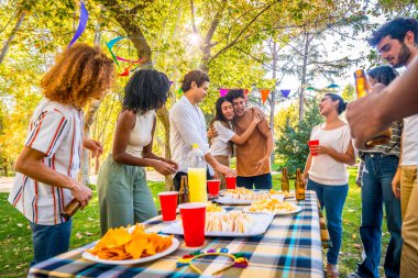 Parktaki piknik partisinde çok ırklı genç arkadaşlara iyi haber verirken kucaklaşan beyaz bir çift.
