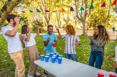 Çeşitli mutlu arkadaşlar kutlama yaparken parkta bira pong oynuyorlar.