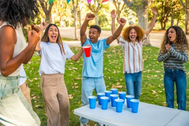Mutlu çoklu etnik arkadaşlar parkta bira pong oynarken silah kaldırıyor.
