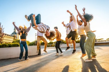 Kıvırcık saçlı bir adam ters takla atıyor. Gün batımında arkadaşlarıyla eğleniyor.