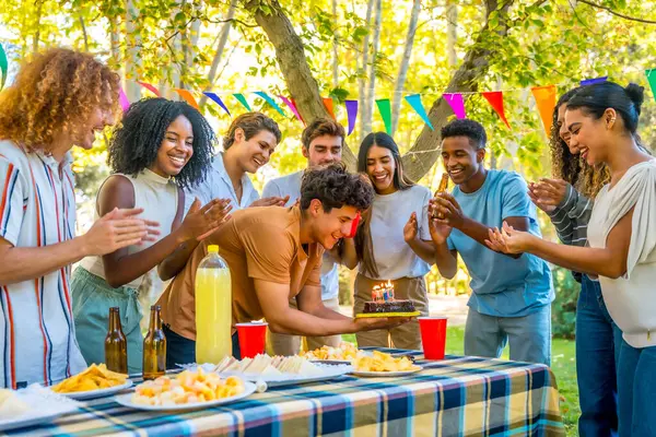 Çok ırklı arkadaşlar parkta piknik yaparken pastalı bir adama doğum günü şarkısı söylüyorlar.