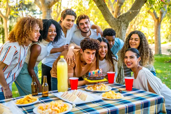 Genç beyaz adam halka açık bir parkta çok ırklı arkadaşlarla piknik yaparken doğum günü pastası mumları üflüyor.